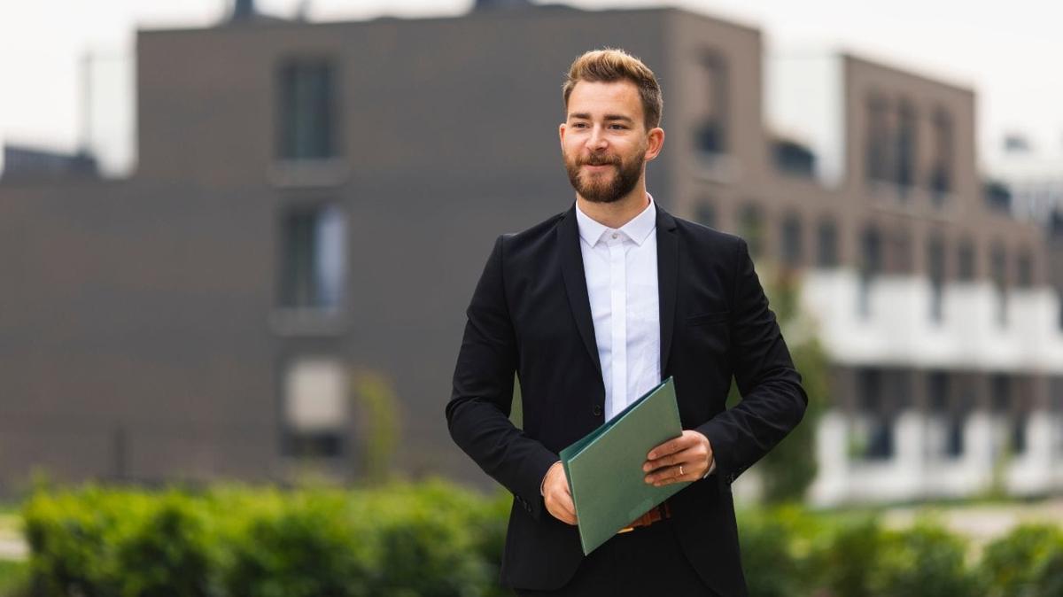 A Realtor in a black suit and a white collared shirt, without a tie, walks outside with a smile, exuding confidence and approachability.