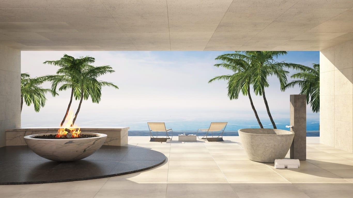 A stone terrace of a luxury residence with a marble fire bowl, a carved stone bathtub and lounge chairs facing palms and the sea.
