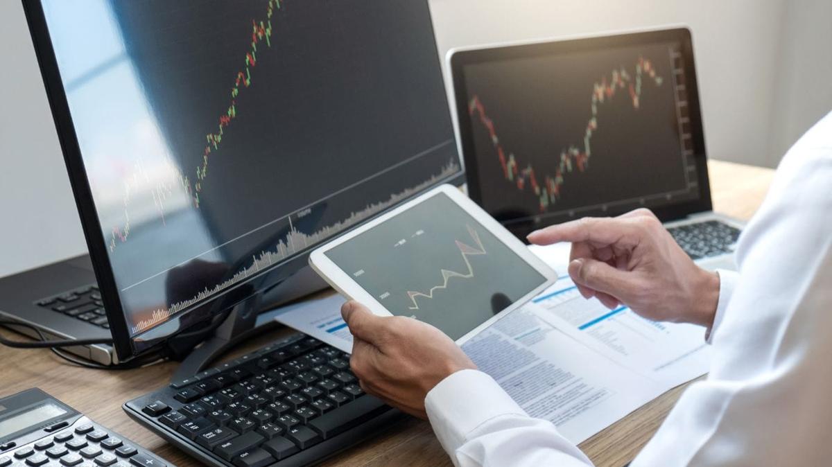 A trader with two screens in front of him displaying stock charts, while holding a tablet that shows a Dividend Growth Stock chart
