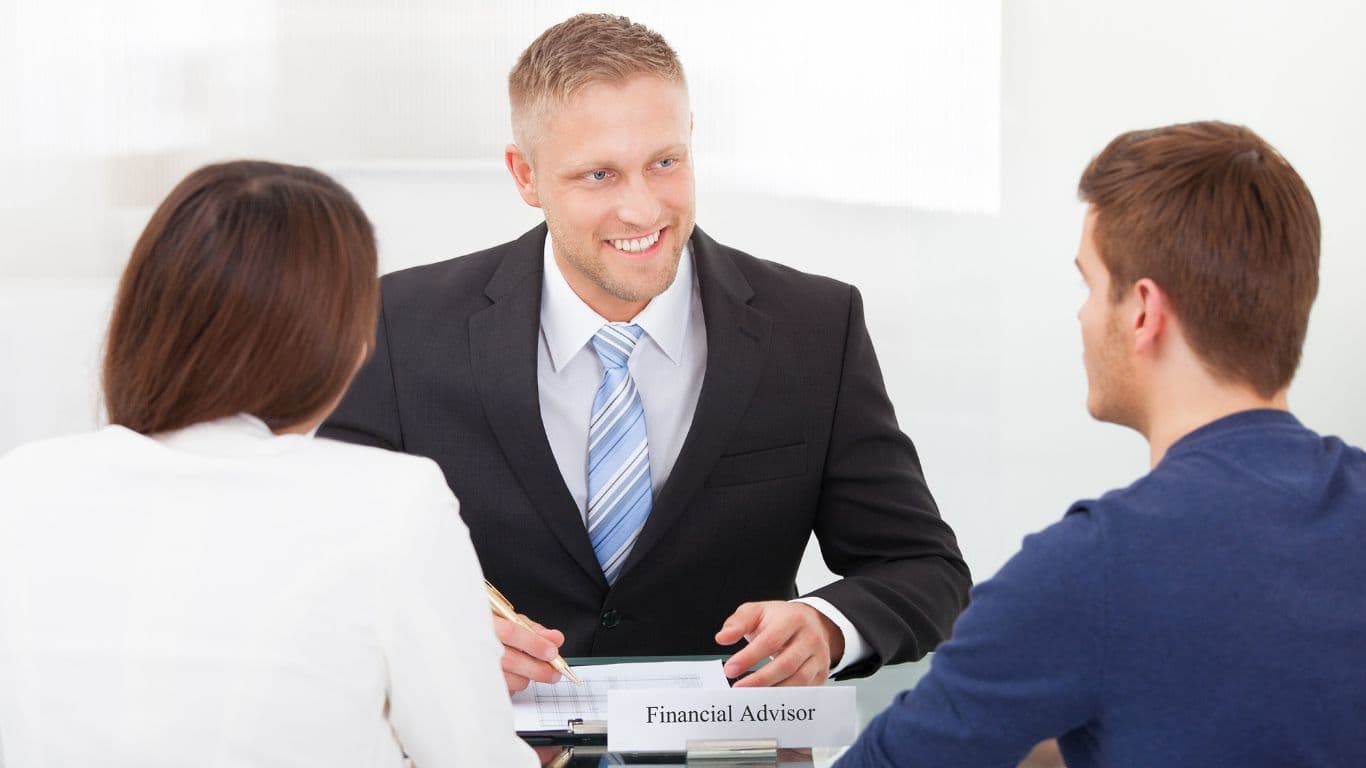 A financial advisor meets with a couple in his office, seated across from him at a desk cluttered with various documents. The advisor smiles warmly at the man while jotting down notes, creating an engaging and friendly atmosphere.