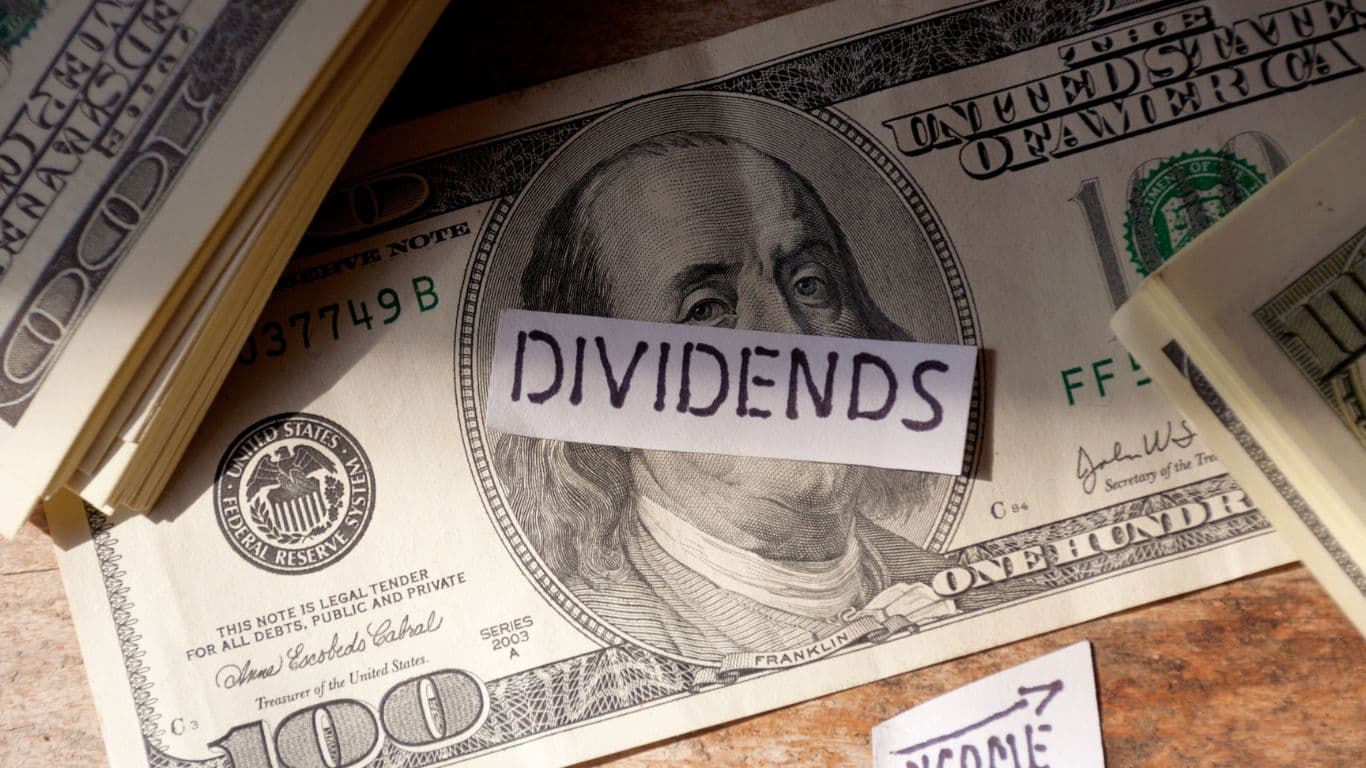 A table with a stack of $100 bills on the left and a single $100 bill in the middle, with a small piece of paper labeled "Dividends" placed in the center of the bill