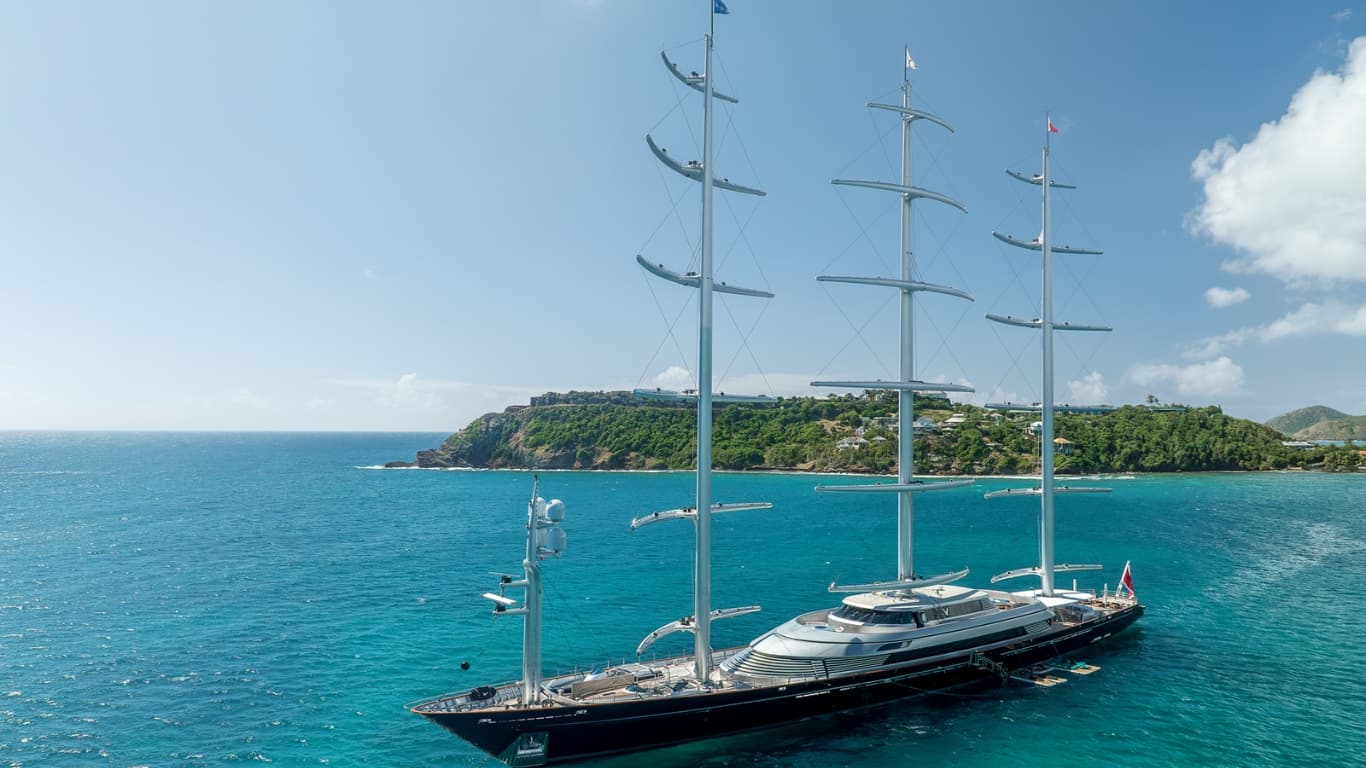 The 88-metre Maltese Falcon at anchor in Caribbean waters, her three rotating carbon-fibre DynaRig masts unmistakable against the headland — the only fully realised proof of Wilhelm Prölss's 1960s rig at sea-going scale.