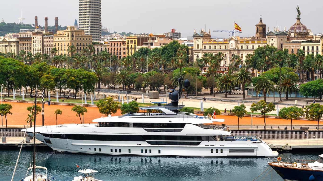 A modern superyacht moored in Barcelona historic port against the Spanish flag and palm-lined waterfront — European yachting in 2026 where compliance structure has become a core component of valuation.