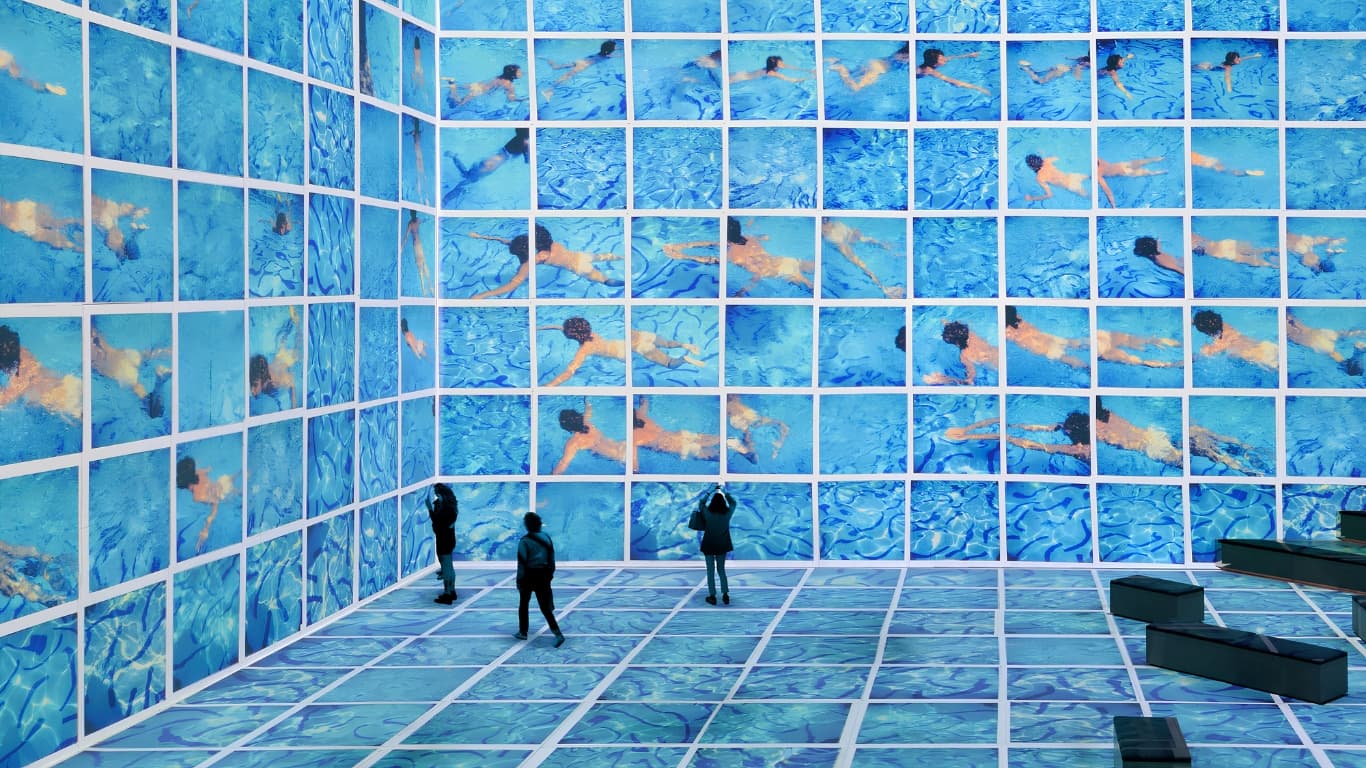 Visitors inside an immersive David Hockney installation, the walls and floor tiled with his blue swimming pool paintings of swimmers.