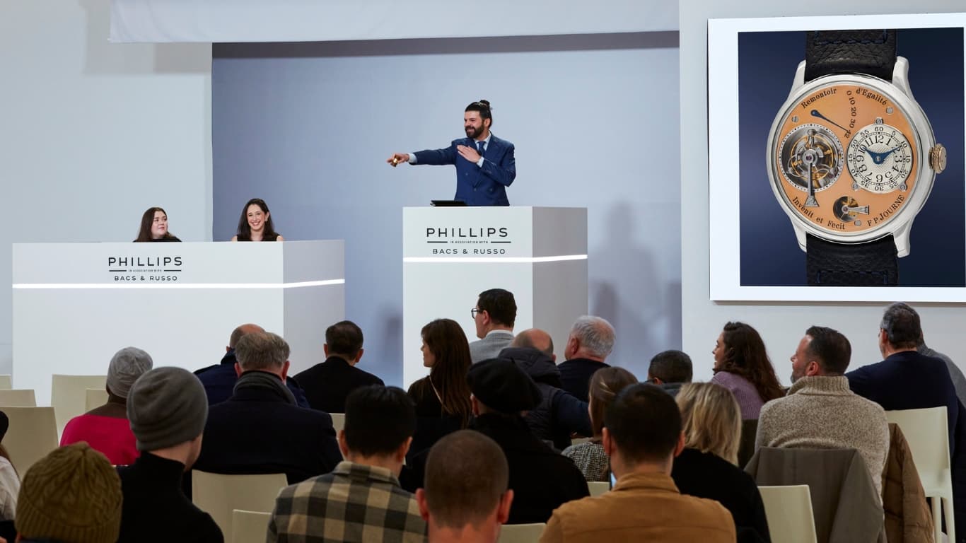 An auctioneer on the rostrum at a Phillips Bacs and Russo watch sale gesturing to bidders, with an F.P. Journe tourbillon shown on the screen alongside.