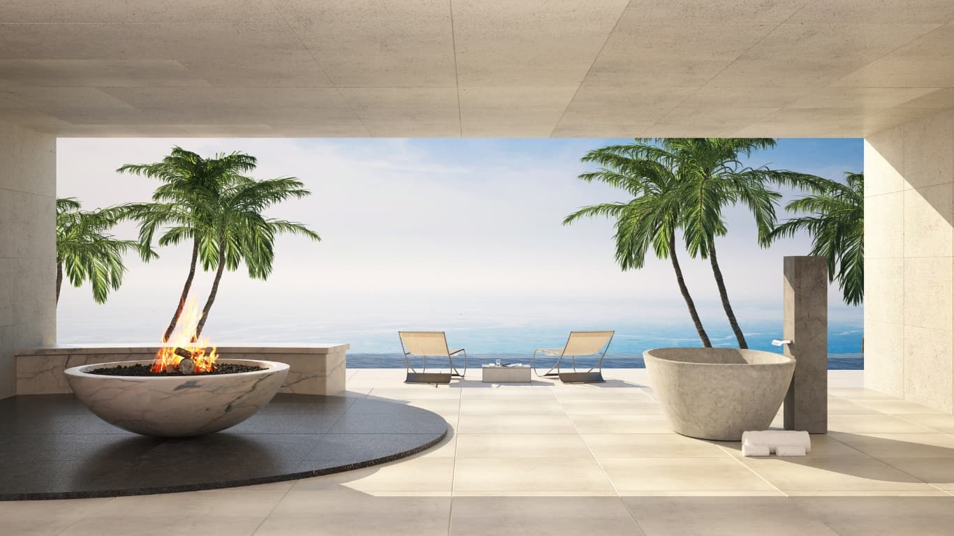 A stone terrace of a luxury residence with a marble fire bowl, a carved stone bathtub and lounge chairs facing palms and the sea.