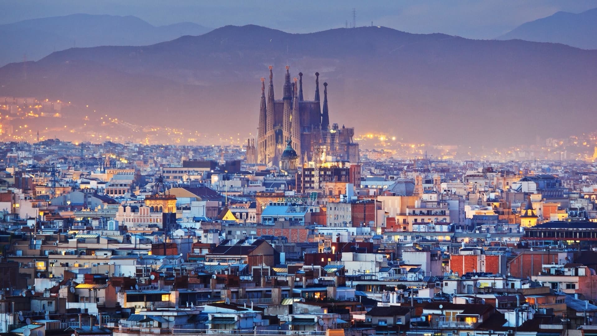 The Barcelona skyline at dusk with the towers of the Sagrada Familia rising above the rooftops and hills behind.