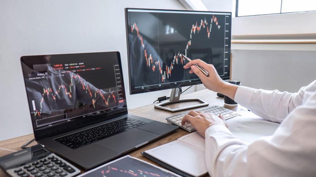 A trader intently examines a stock chart, pointing at key details with his pen. To his left, a laptop displays another open chart, providing additional market data as he analyzes both screens with focused precision.