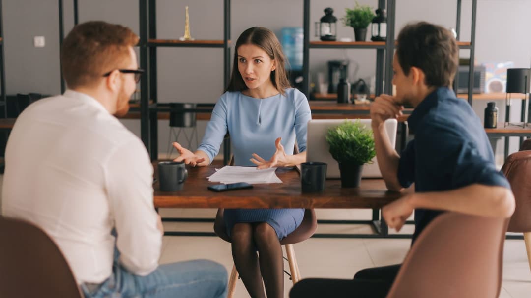 A dual agent is seated at a desk with both the buyer and seller of a house, discussing the final details of the transaction. The atmosphere is collaborative as they clarify terms and address any last-minute questions