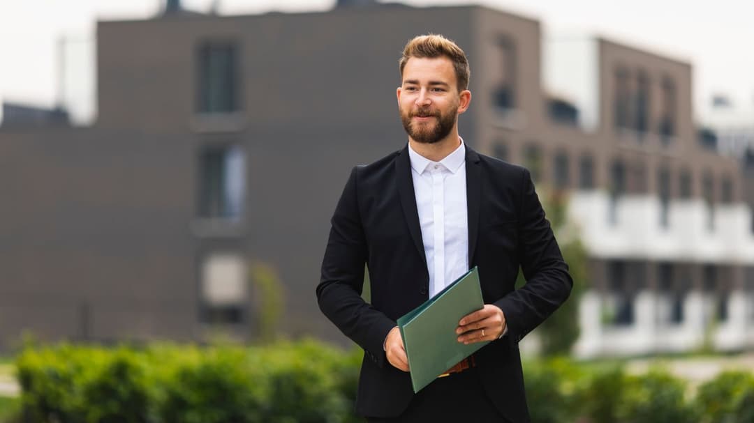 A Realtor in a black suit and a white collared shirt, without a tie, walks outside with a smile, exuding confidence and approachability.
