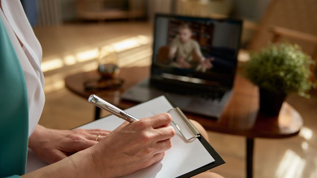 A female psychologist on a video call with a male client. As he speaks, she listens while jotting down notes on a piece of paper.