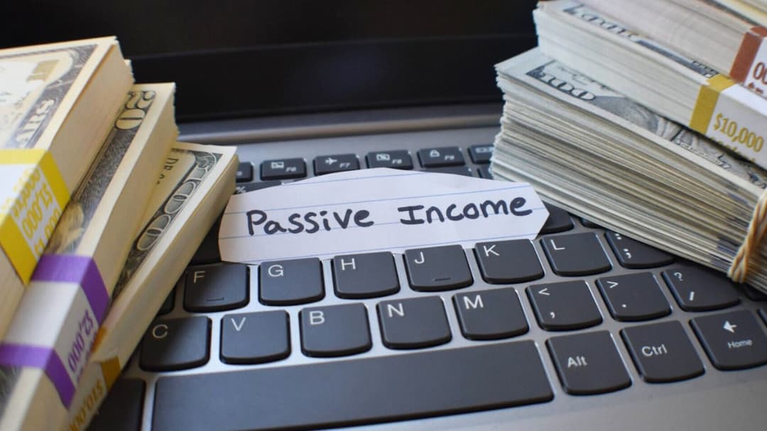 A laptop with stacks of dollar bills on either side of the keyboard and a piece of paper in the center labeled "Passive Income"