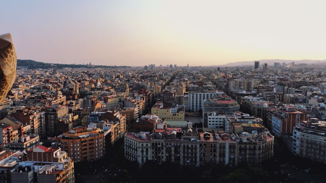 Rooftop view over the dense residential blocks of Barcelona at dusk, the Eixample grid stretching toward the hills.
