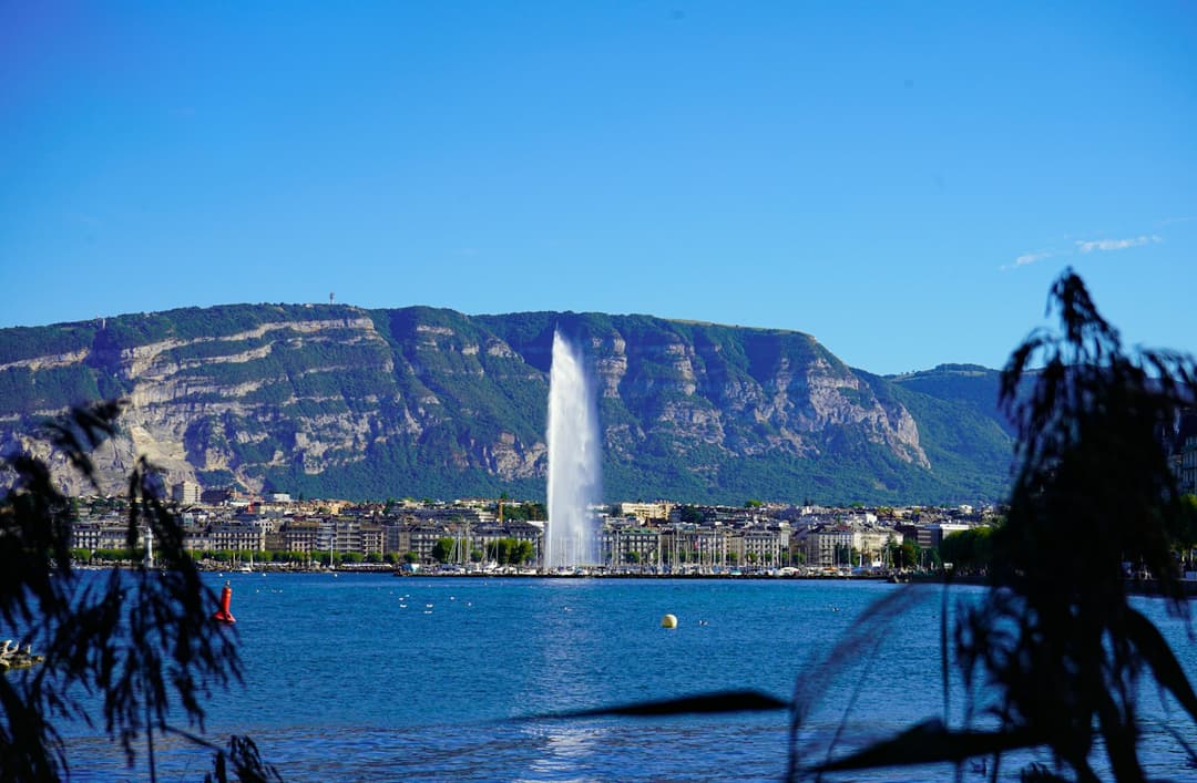 The Jet d'Eau fountain rising over Lake Geneva with the city lakefront and hills behind, in the city where the article opens.