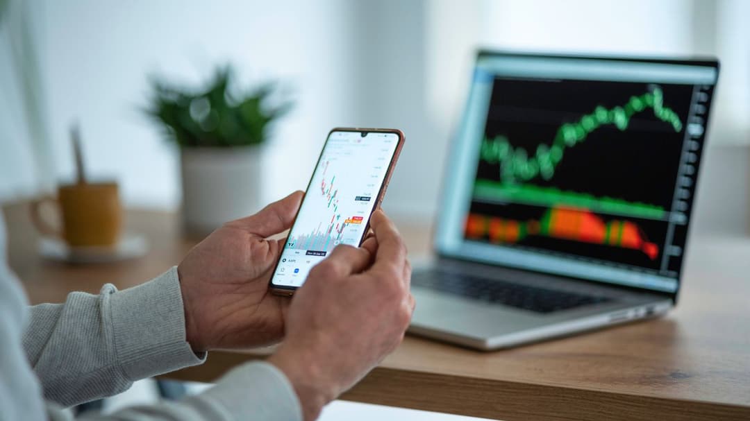 A trader is setting up a Couch-Potato Portfolio on his phone, while in front of him, a table holds a laptop displaying an active trading chart.