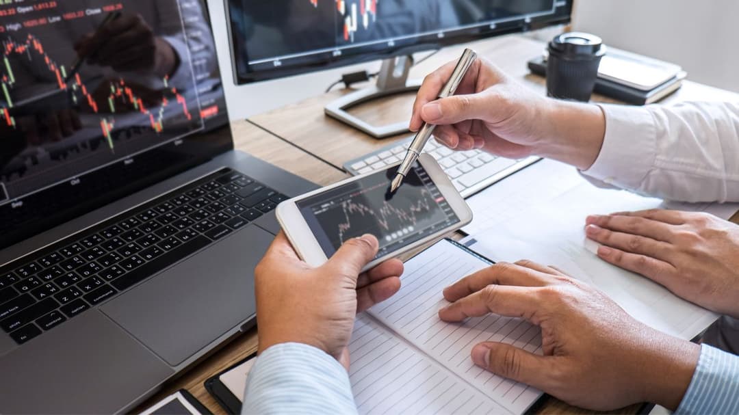 Two men are in front of a laptop and monitor displaying charts. One man holds a phone showing a chart, while the other points at it with a pen, explaining the types of indexes that require rebalancing.