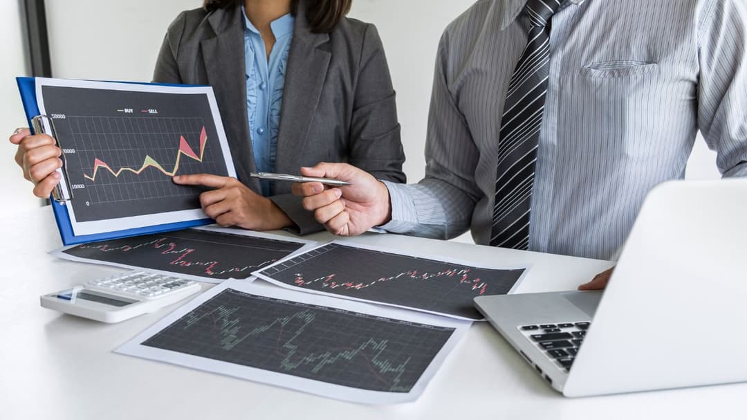 An investor is explaining the concept of a Couch-Potato Portfolio to a woman who holds a photo of a trading chart. As he points to a specific area on the chart with his pen, she mirrors his focus by pointing at the same spot with her finger, fully engaged in the discussion.