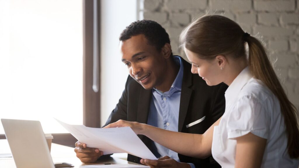 A financial analyst and a client sit at a desk, with a laptop nearby. The client holds a piece of paper, and the analyst points to specific details on it as they discuss its contents, focusing on financial strategies and insights.