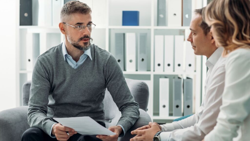 A financial advisor meets with two clients in a cozy office setting. The clients are seated on a comfortable sofa, while the advisor sits across from them in an armchair. Together, they engage in a thoughtful discussion, reviewing financial options and strategies