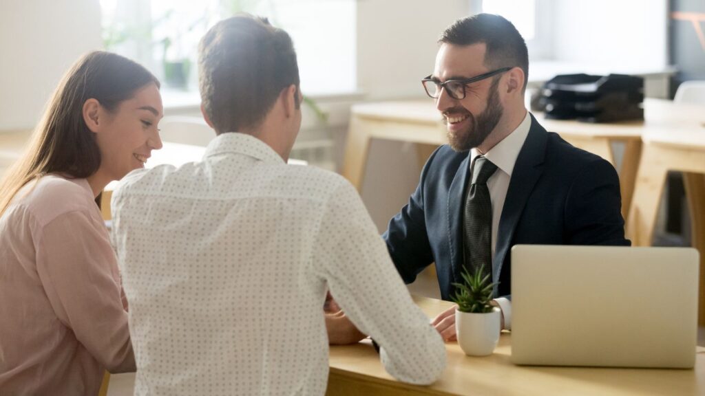 A financial advisor sits at a desk with two clients, engaged in conversation. The advisor's laptop is open to the side, creating a professional and collaborative atmosphere.