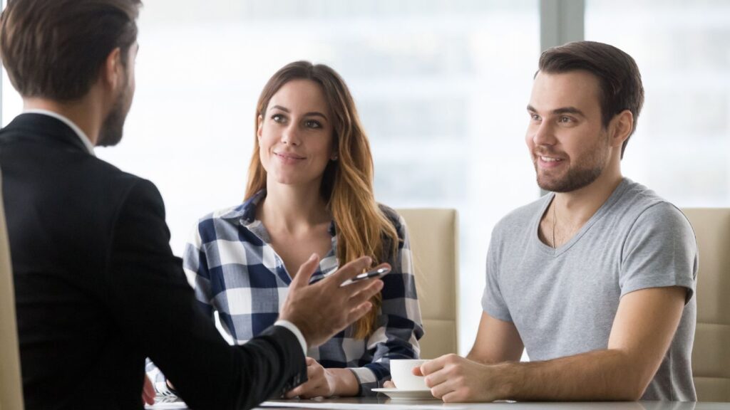 A financial advisor meets with a couple in his office. The man holds up a coffee cup, attentively listening, while the woman smiles, engaged in the conversation. The advisor shares insights, creating a warm and collaborative atmosphere as they discuss their financial plans together.