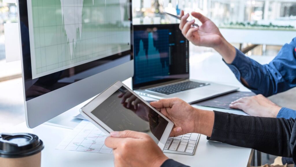 Two investors are seated in front of a monitor displaying a trading chart, engaged in discussion. One points at the screen with a pen, while the other holds a tablet. On the desk, a laptop also displays a trading chart.
