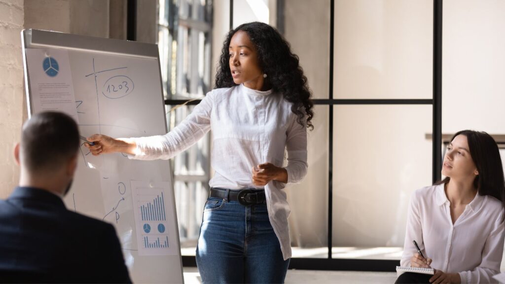 A woman stands in front of a whiteboard, talking and pointing at numbers she previously wrote, while two people in front of her take notes attentively