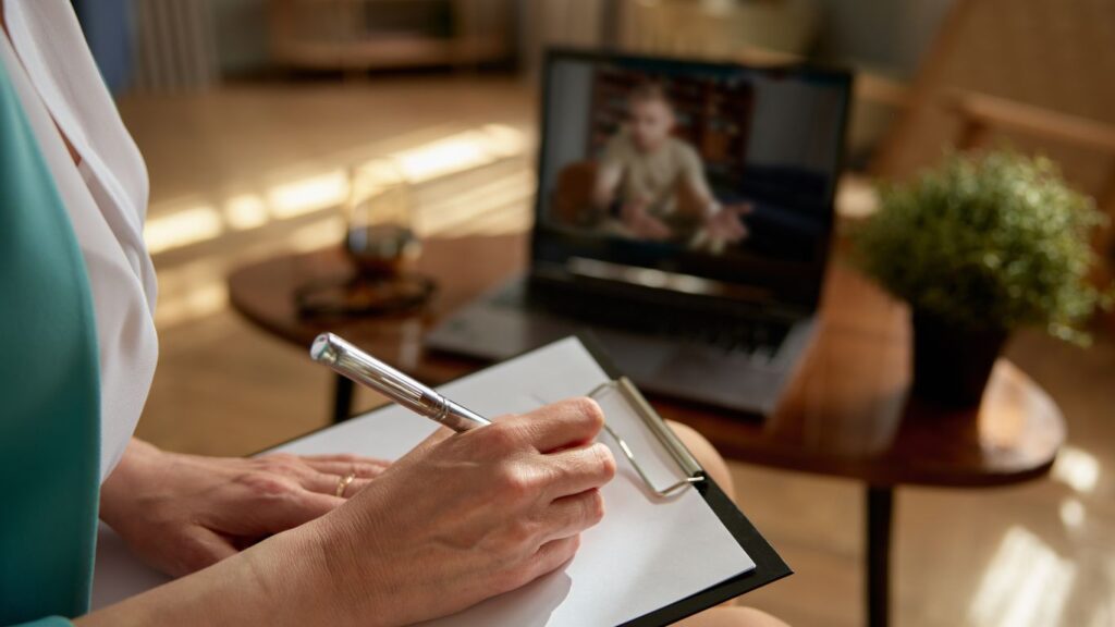 A female psychologist on a video call with a male client. As he speaks, she listens while jotting down notes on a piece of paper.