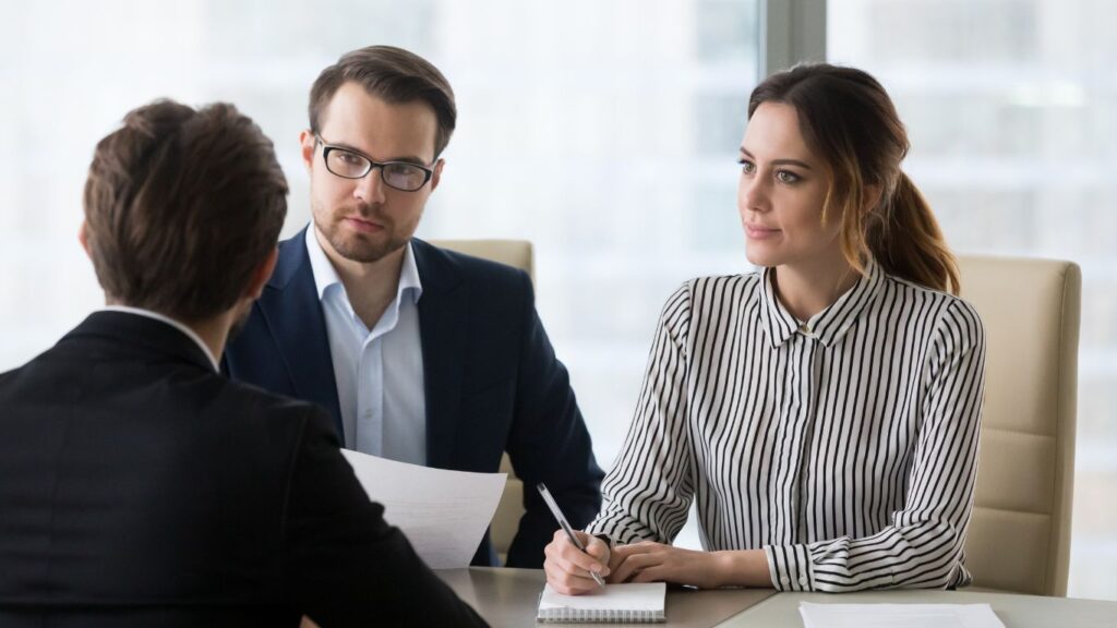 An asset manager addresses a male and a female client standing before him, both attentively listening and taking notes as he speaks