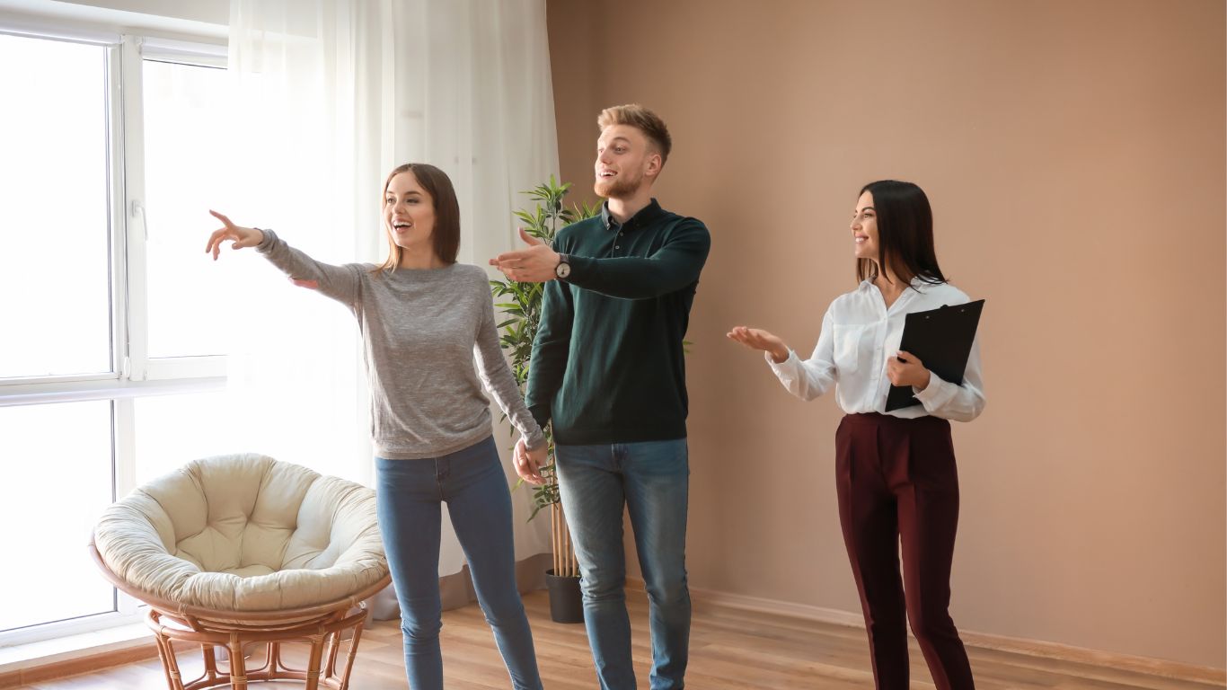 A smiling real estate agent shows an excited young couple around a house. The agent, holding a black folder, gestures confidently. The woman from the couple, holding her partner's hand, eagerly points out a feature of the home, radiating joy and excitement.