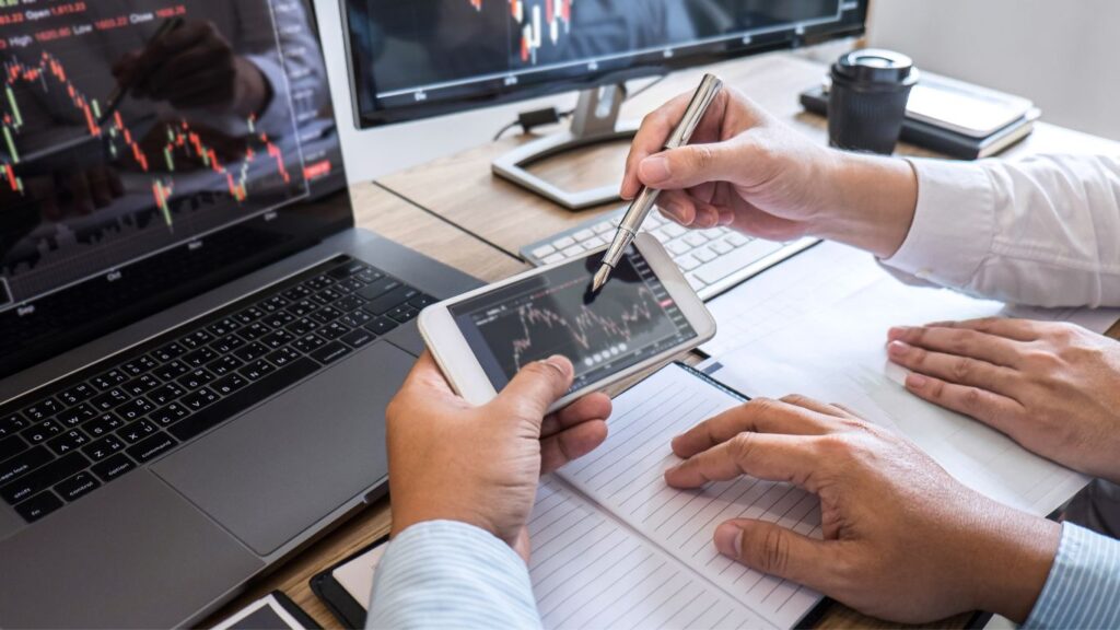 Two men are in front of a laptop and monitor displaying charts. One man holds a phone showing a chart, while the other points at it with a pen, explaining the types of indexes that require rebalancing.
